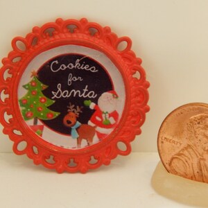 May include: A red, ornate, circular Christmas ornament with the text "Cookies for Santa." The ornament features a festive design with a Christmas tree, Santa Claus, and a reindeer. A penny is next to the ornament.