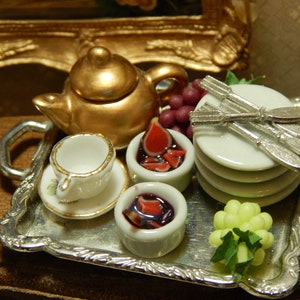 May include: A silver serving tray with a gold teapot, a small white teacup, two small white bowls with red fruit preserves, a stack of white plates, a silver knife and fork, and a bunch of green grapes.