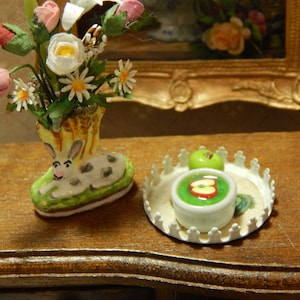 May include: A white ceramic bowl with green apple slices and a green liquid inside, on a white tray with a decorative border. A green apple sits on the tray next to the bowl. The tray is on a wooden surface. A ceramic boot with a rabbit face and flowers in it is in the background.