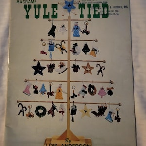 Op de afbeelding: Een vintage macramé knutselinstructieboekje getiteld "Yule Tied" met een kerstboomontwerp. De omslag toont een boom versierd met diverse macramé ornamenten in wit, blauw en goud. De naam van de auteur, Lois Anderson, staat onderaan.