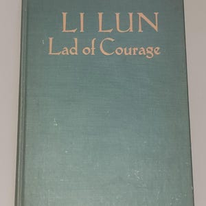 May include: A vintage book titled "LI LUN Lad of Courage." The book has a teal cover with the title and subtitle in a contrasting light brown color. The book's cover is slightly worn, suggesting its age.