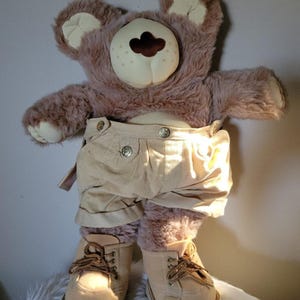 May include: A plush teddy bear wearing tan corduroy shorts and matching boots with brown laces. The bear has a light brown face and ears, and a darker brown fur body. The bear is standing on a white, fluffy surface.
