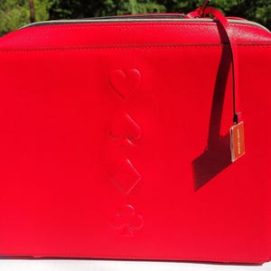 Può includere: Borsa rettangolare rosso brillante con simboli di carte da gioco in rilievo: cuori, picche, quadri e fiori. La borsa ha una chiusura a zip e un piccolo cartellino attaccato con un nastro rosso.