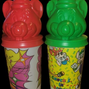May include: Two vintage plastic cups with lids, one red and one green, featuring cartoon character faces. The cups have colorful graphics, including the text "Chuck E. in Action!" and retro designs. The cups are white at the bottom.