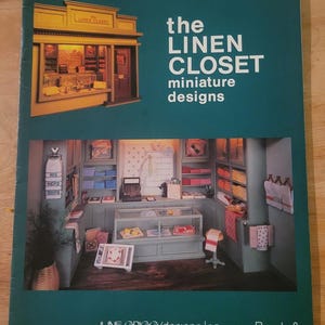 Op de afbeelding: Een turquoise boekomslag met de titel "the LINEN CLOSET miniature designs". De omslag toont een miniatuurwinkelinterieur met planken vol linnen en een etalage. De prijs is €368.