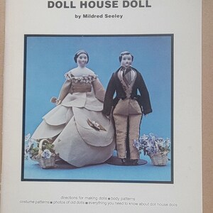May include: The cover of a book titled "THE DOLL HOUSE DOLL" by Mildred Seeley. The cover features a framed image of two doll house dolls, a woman in a white dress and a man in a black suit. The cover also includes text about doll making.