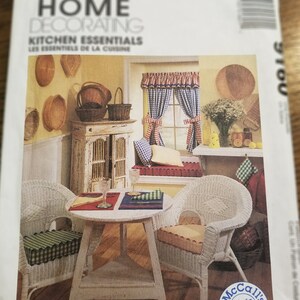 May include: A McCall's Home Decorating pattern cover featuring a kitchen scene. The image shows a white wicker table and chairs, a window with gingham curtains, and a cabinet. The text reads "Kitchen Essentials".
