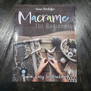 May include: A softcover book titled Macrame for Beginners by Vivien Romhoffer with the subtitle Super Easy Instructions. The cover features a photograph of various macrame projects, yarn balls, and hands working on a piece of white cord art.
