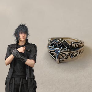 May include: Silver-toned ring with an ornate design featuring a central blue gemstone. The ring has intricate detailing and a stylized, possibly fantasy-inspired aesthetic. The ring is displayed on a neutral background.