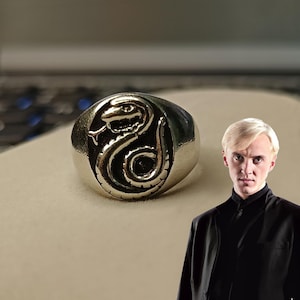 Pode incluir: Anel prateado com um design detalhado de cobra. O anel apresenta um emblema de cobra em relevo com escamas intrincadas e um corpo curvo. A cobra est&aacute; centrada em uma banda redonda e polida. &Eacute; uma joia.