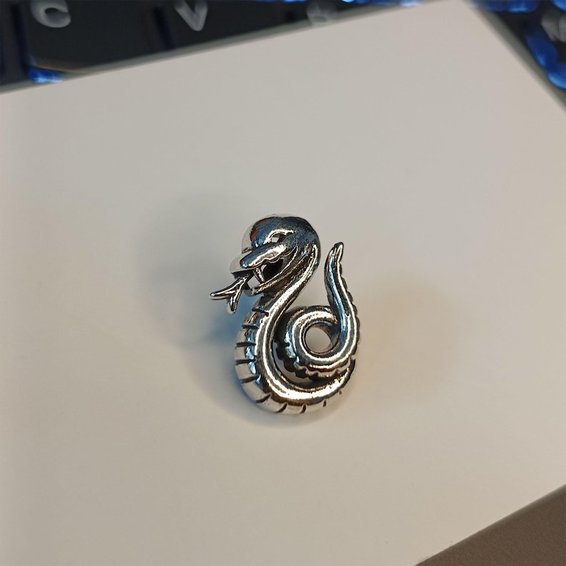 May include: A silver-colored snake pendant with intricate detailing. The snake is coiled, with its head raised and tongue extended. The pendant has a polished finish, reflecting light. The design is a stylized representation of a snake.
