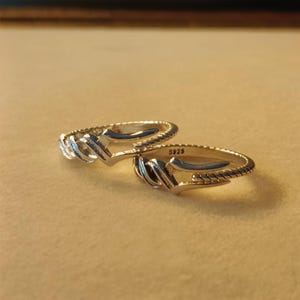May include: Two silver rings with a unique design. Each ring features a textured band and a stylized arrow-like shape. The rings are marked with "S925". The rings are displayed on a neutral background.