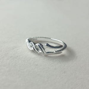 May include: A silver ring with a unique design. The ring features a twisted, rope-like pattern in the center, with a smooth, polished finish. The band has a textured, beaded edge. The ring is shown on a white surface.