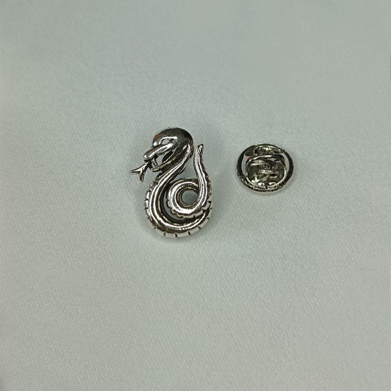 May include: A silver-colored snake lapel pin with intricate detailing, coiled in a stylized design. The pin features a separate backing piece for secure attachment. The snake's head is raised, with a forked tongue visible.