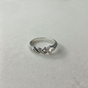 May include: A silver ring with a braided design. The ring features a textured band and an interwoven pattern in the center. The ring is displayed on a white surface.