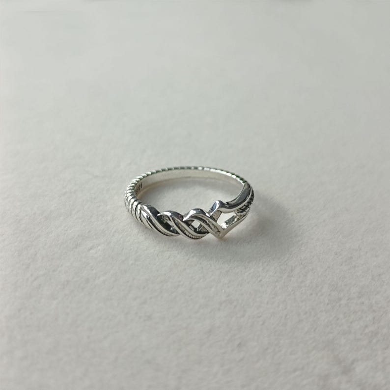 May include: A silver ring with a braided design. The ring features a textured band and an interwoven pattern in the center. The ring is displayed on a white surface.