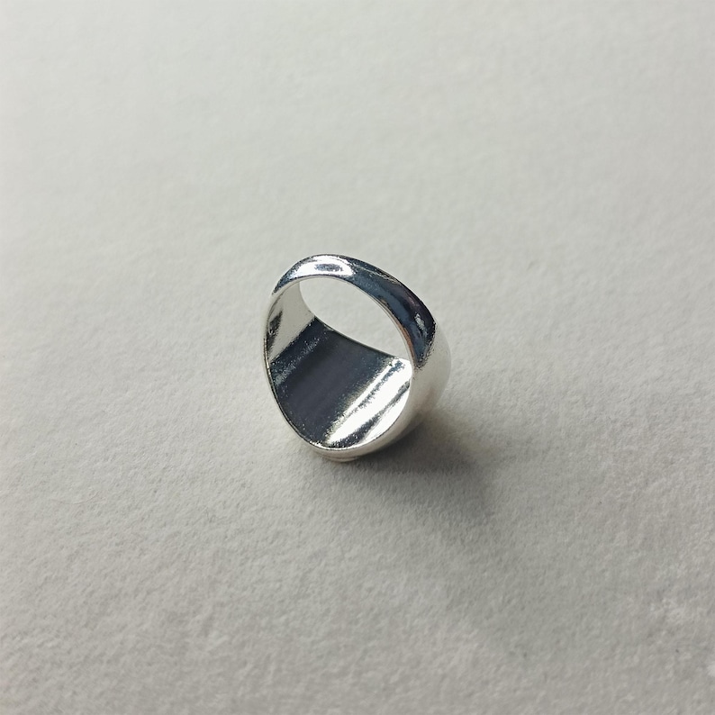 Pode incluir: Um anel prateado brilhante com um design liso e arredondado. O anel tem um interior plano e retangular e um acabamento polido, refletindo a luz. Uma joia simples e elegante.