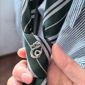 May include: A green and white striped tie with a silver snake pin. The tie is knotted and held in place by the pin, which features a detailed snake design. The tie is made of a smooth fabric.