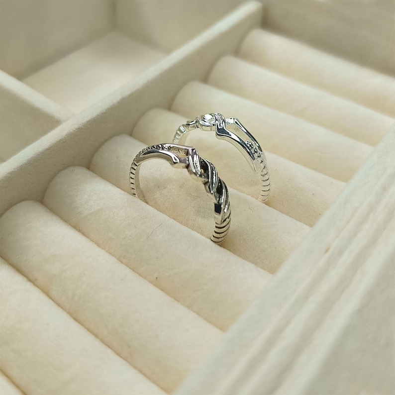 May include: Two silver rings with intricate designs rest in a cream-colored jewelry box. One ring features a twisted rope design, while the other has a delicate, openwork pattern with small embellishments. The rings are displayed on a soft, velvety surface.