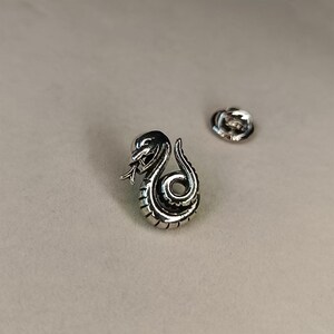 May include: A silver-colored snake pin with intricate detailing, coiled in a striking pose with its tongue extended. The pin is accompanied by a separate pin back, suggesting it's designed to be attached to clothing or accessories.