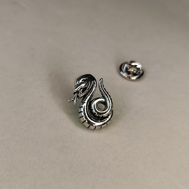 May include: A silver-colored snake pin with intricate detailing, coiled in a striking pose with its tongue extended. The pin is accompanied by a separate pin back, suggesting it's designed to be attached to clothing or accessories.
