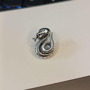May include: A silver-colored snake pendant with intricate detailing. The snake is coiled, with its head raised and tongue extended. The pendant is likely made of metal and has a polished finish. The design is detailed and realistic.