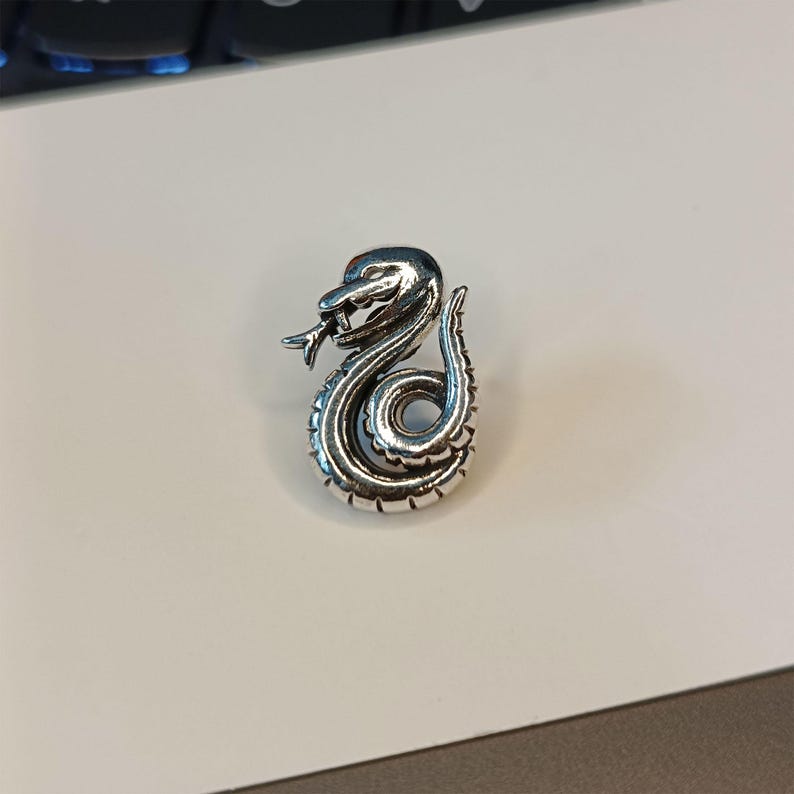 May include: A silver-colored snake pendant with intricate detailing. The snake is coiled, with its head raised and tongue extended. The pendant is likely made of metal and has a polished finish. The design is detailed and realistic.