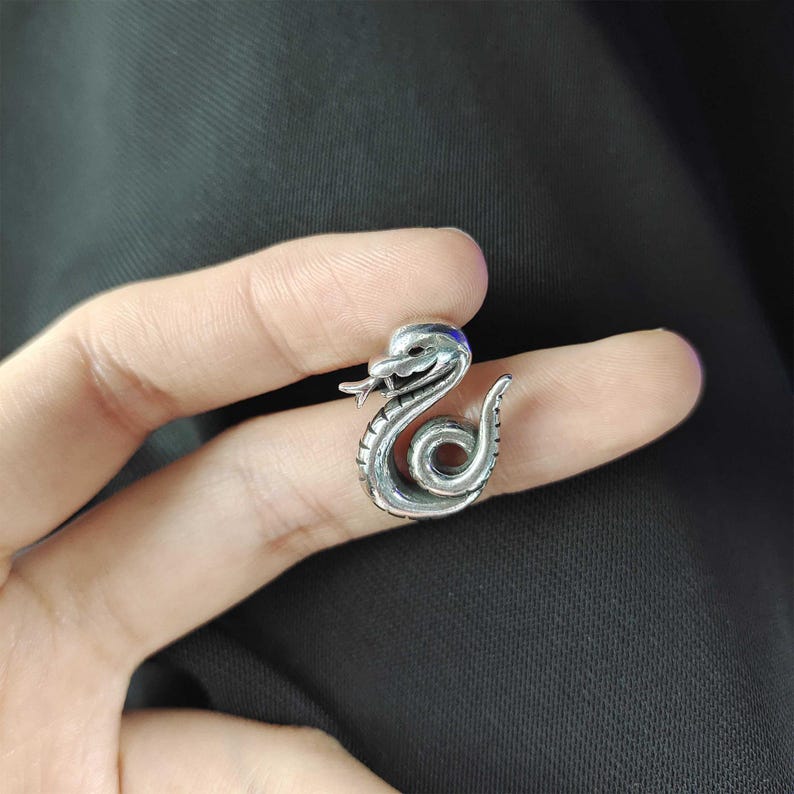 May include: A silver-toned snake-shaped ring with intricate detailing. The snake is coiled with its head raised, tongue extended, and eyes detailed. The ring is a unique piece of jewelry.