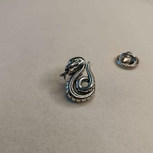 May include: A silver-colored snake pin with intricate detailing, featuring a coiled body and a forked tongue. The pin is accompanied by a separate backing piece. The snake design is a symbol of transformation and healing.