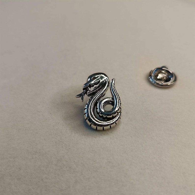 May include: A silver-colored snake pin with intricate detailing, featuring a coiled body and a forked tongue. The pin is accompanied by a separate backing piece. The snake design is a symbol of transformation and healing.
