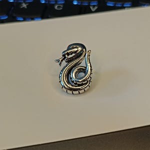 May include: A silver-colored snake figurine in a coiled position, with its head raised and tongue extended. The snake has detailed scales and a slightly open mouth. The figurine is on a white surface.