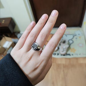 Pode incluir: Um anel prateado com um design preto estilizado na parte superior. O anel &eacute; usado num dedo, mostrando os seus detalhes. O design parece ser um redemoinho ou s&iacute;mbolo abstrato, adicionando um toque &uacute;nico &agrave; joia.