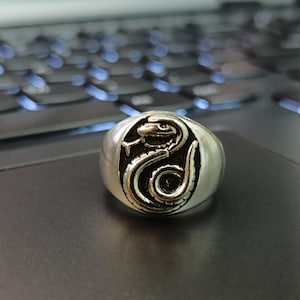 Pode incluir: Um anel prateado com um design detalhado de cobra. A cobra est&aacute; enrolada dentro de um oval escuro, criando um contraste marcante. O anel &eacute; provavelmente feito de metal e tem um acabamento polido, adequado para joias.