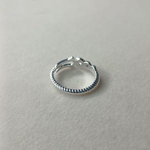 May include: A silver ring with a decorative design. The ring features a textured band and a stylized openwork design on the top. The ring is shown on a neutral background.