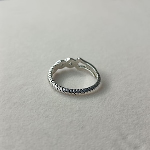 May include: A silver ring with a textured, rope-like band. The ring features three small, diamond-shaped accents at the top. The ring is made of a shiny, metallic material.