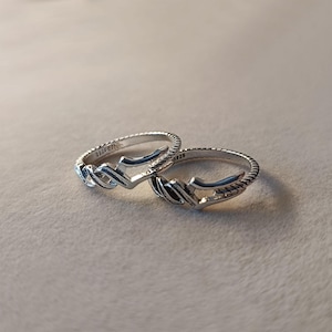 May include: Two silver rings with a textured band and a stylized arrow design. The rings are made of a shiny metal and are displayed on a neutral background. The rings are marked with "S925".
