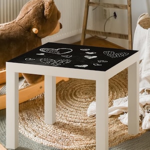 Può includere: Un tavolo quadrato bianco con un piano nero con disegni a tratto bianco di cuori e altre forme. Il tavolo è su un tappeto rotondo di iuta. Un grande cavallo a dondolo a forma di orsacchiotto marrone è sullo sfondo, insieme a una scala di legno e un quadro incorniciato.