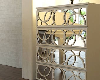 Può includere: Una cassettiera a quattro cassetti con finitura color crema chiaro. Ogni frontale del cassetto presenta un rivestimento decorativo di cerchi intrecciati in una tonalità crema abbinata, su uno sfondo a specchio che riflette l'ambiente.