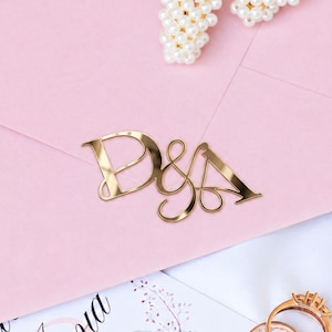 Pode incluir: Um envelope rosa claro selado com um monograma metálico dourado com as letras D e A. Dois ganchos de cabelo com pérolas repousam sobre o envelope, juntamente com dois anéis dourados e um convite com estampa floral e caligrafia.
