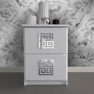 Può includere: Comodino bianco con due cassetti e tiretti con motivo geometrico argentato. Il comodino è su uno sfondo di carta da parati floreale bianca.