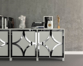 Può includere: Un mobile bianco moderno con disegni geometrici a specchio sulle ante. Il mobile è sormontato da oggetti decorativi, tra cui libri, un busto, una foto incorniciata, un vaso e una scultura dorata. Lo sfondo è un muro grigio.