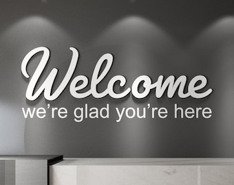 May include: A modern reception area with a gray wall featuring the word "Welcome" in a white, cursive font, followed by "we're glad you're here." A marble-patterned counter and a small white vase with a plant are visible.
