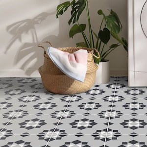 May include: A white washing machine with a basket of laundry inside, sitting on a gray and white patterned tile floor. A potted plant and a basket of towels are on a shelf above the washing machine.
