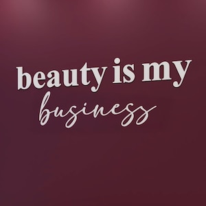 Könnte beinhalten: Innenraum eines Schönheitssalons mit einer weinroten Wand mit dem weißen Text "beauty is my business". Ein ovaler Spiegel und ein runder Spiegel sind sichtbar. Unter dem Text befindet sich eine Bank mit brauner Oberseite und weißem Sockel.
