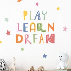 Peut inclure: Décoration murale avec les mots "PLAY LEARN DREAM" en lettres colorées et des étoiles. Un ours blanc en guise de lampe, des jouets en bois et un meuble de rangement créent une ambiance enfantine.