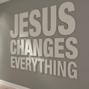 May include: White 3D lettering on a grey wall reads "JESUS CHANGES EVERYTHING." The words are arranged in three lines, with "JESUS" at the top, "CHANGES" in the middle, and "EVERYTHING" at the bottom. The wall is painted a solid grey colour.
