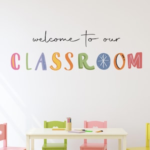 May include: A classroom scene with a white table and colorful chairs. The chairs are pink, green, and yellow. The wall has the text "welcome to our CLASSROOM" in black and colorful letters. A rug is on the floor.