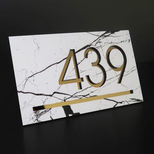 May include: A rectangular address plaque with a white marble-like background and black and gold accents. The numbers "439" are prominently displayed in gold and black, with a gold line beneath them. The plaque is angled against a dark background.