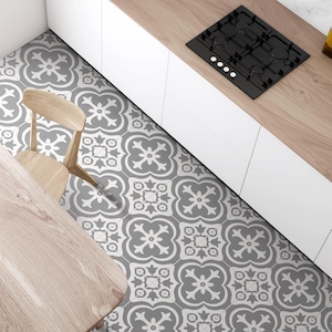 Peut inclure: Un sol de cuisine composé de carreaux carrés avec un motif floral gris et blanc répétitif. La cuisine comprend des armoires blanches, un plan de travail en bois clair, une plaque de cuisson noire, une chaise et un coin de table en bois.