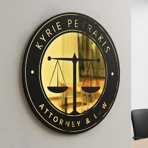 Può includere: Un cartello rotondo nero e oro con la scritta "KYRIE PETRAKIS ATTORNEY & LAW". Il cartello presenta un centro dorato con un simbolo della bilancia della giustizia. Il cartello è montato su una parete bianca.